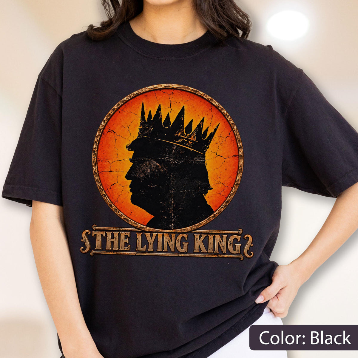 May include: A black t-shirt featuring a circular graphic with a distressed orange and yellow background. Inside the circle is a black silhouette of a person wearing a crown. Below the circle, the text THE LYING KING is printed in a gold font.