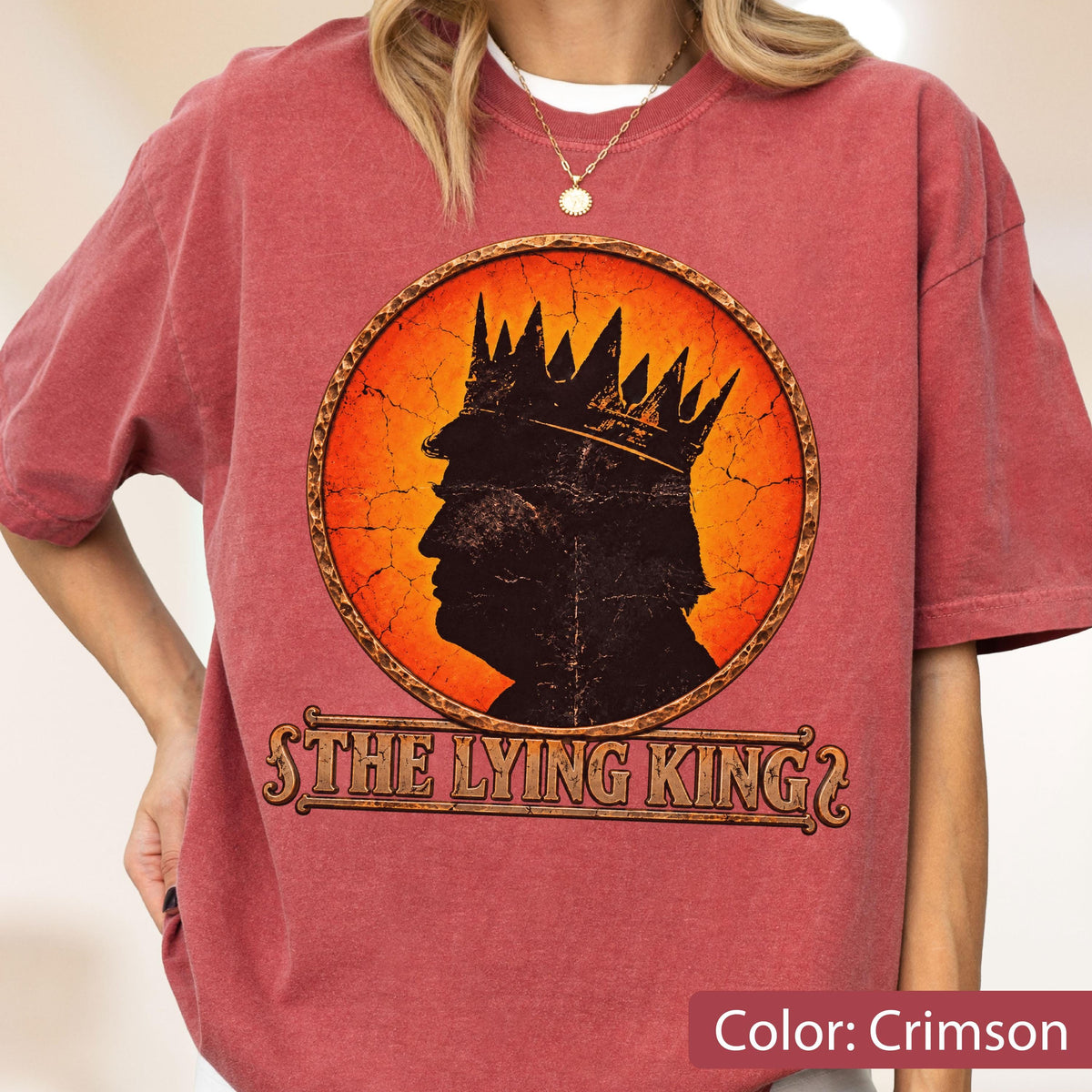 May include: A crimson short-sleeve t-shirt featuring a circular graphic with a black silhouette of a person wearing a crown against an orange cracked background. Below the circle, the text reads THE LYING KING in a stylized, weathered font.