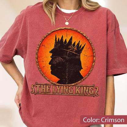 May include: A crimson short-sleeve t-shirt featuring a circular graphic with a black silhouette of a person wearing a crown against an orange cracked background. Below the circle, the text reads THE LYING KING in a stylized, weathered font.