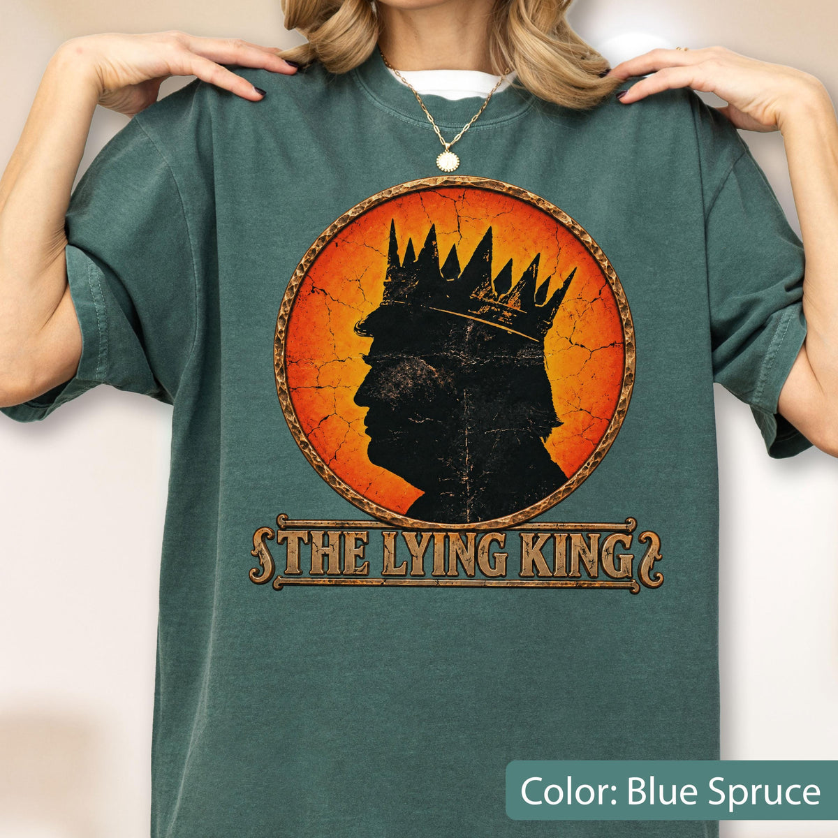 May include: A blue spruce t-shirt featuring a circular graphic with a distressed orange background. The graphic displays a black silhouette of a person wearing a crown. Below the circle, the text reads THE LYING KING in a stylized font.