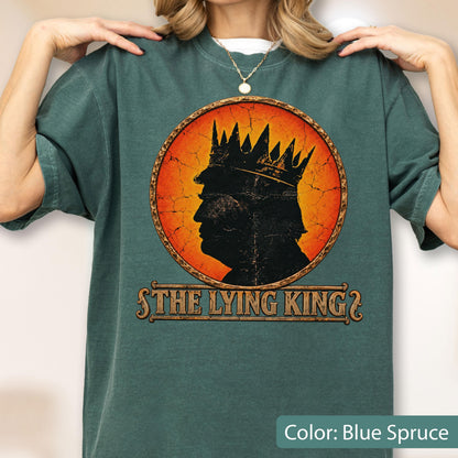May include: A blue spruce t-shirt featuring a circular graphic with a distressed orange background. The graphic displays a black silhouette of a person wearing a crown. Below the circle, the text reads THE LYING KING in a stylized font.
