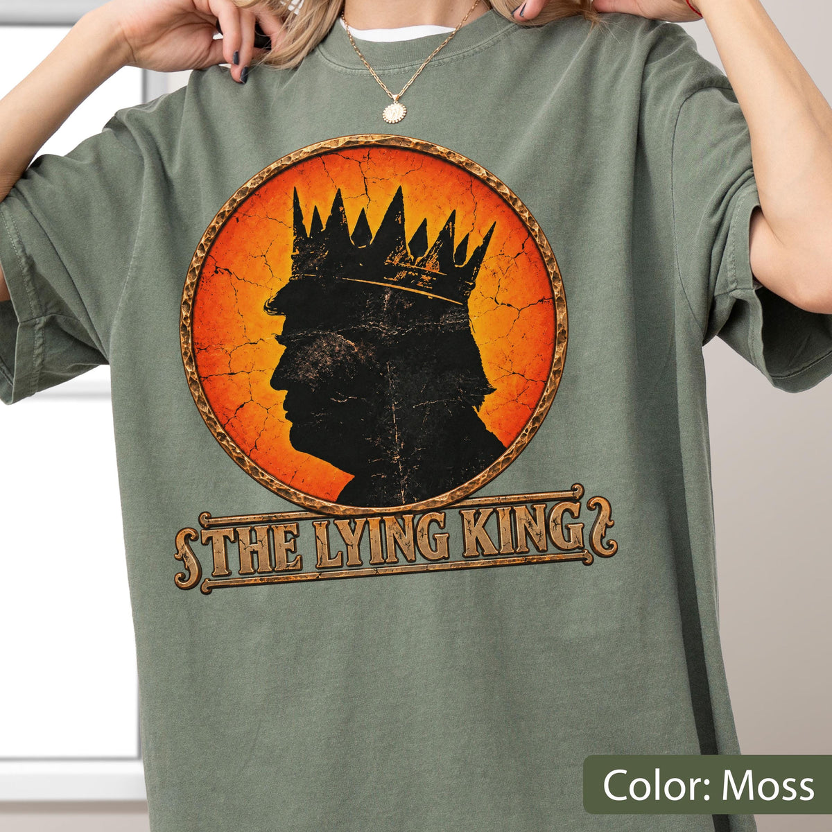 May include: A moss green t-shirt featuring a circular graphic with a distressed orange background. Inside the circle is a black silhouette of a person wearing a crown. Below the graphic, the text reads THE LYING KING in a stylized font.