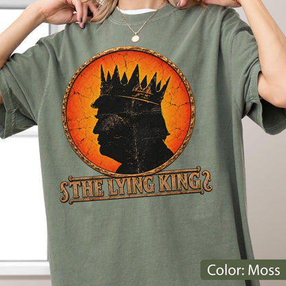 May include: A moss green t-shirt featuring a circular graphic with a distressed orange background. Inside the circle is a black silhouette of a person wearing a crown. Below the graphic, the text reads THE LYING KING in a stylized font.