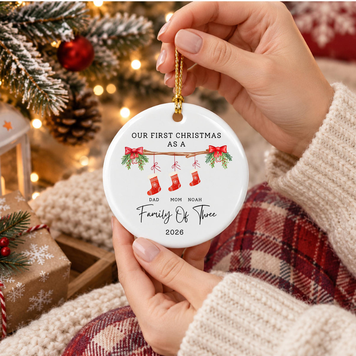 May include: A round white ceramic Christmas ornament featuring a printed illustration of a branch with three hanging red stockings. The text reads: OUR FIRST CHRISTMAS AS A FAMILY OF THREE, DAD, MOM, NOAH, and 2026. It includes a gold hanging cord.