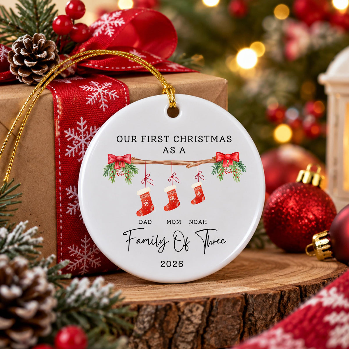 May include: A round white ceramic Christmas ornament featuring a graphic of a wooden branch with three red stockings hanging from it. The text reads: OUR FIRST CHRISTMAS AS A, DAD, MOM, NOAH, Family Of Three, and 2026, with red bows.