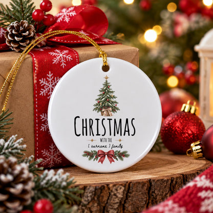 May include: A round white ceramic ornament featuring a green Christmas tree illustration, the text "CHRISTMAS WITH THE [ surname ] family" in black, and a red bow graphic with pine branches at the bottom, suspended by a gold string.