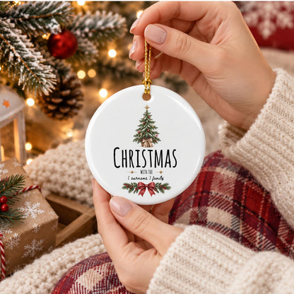 May include: A round white ceramic ornament featuring a printed illustration of a green Christmas tree with a gold star, a red bow, and the text "CHRISTMAS WITH THE [ SURNAME ] family" above a decorative pine branch and red ribbon graphic.
