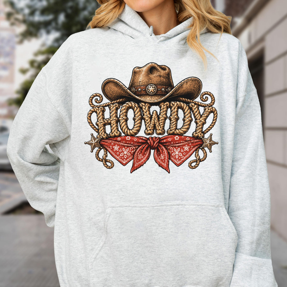 May include: A light gray hooded sweatshirt featuring a graphic print of a brown cowboy hat, a red patterned bandana, and the word HOWDY written in a rope-style font. The design includes two small star icons on the left and right sides.