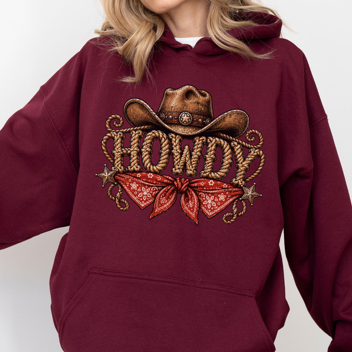 May include: A maroon hooded sweatshirt featuring a graphic print of a brown cowboy hat, the word HOWDY written in a rope-style font, a red bandana with a white floral pattern, and two small metallic-colored stars on the sides of the design.