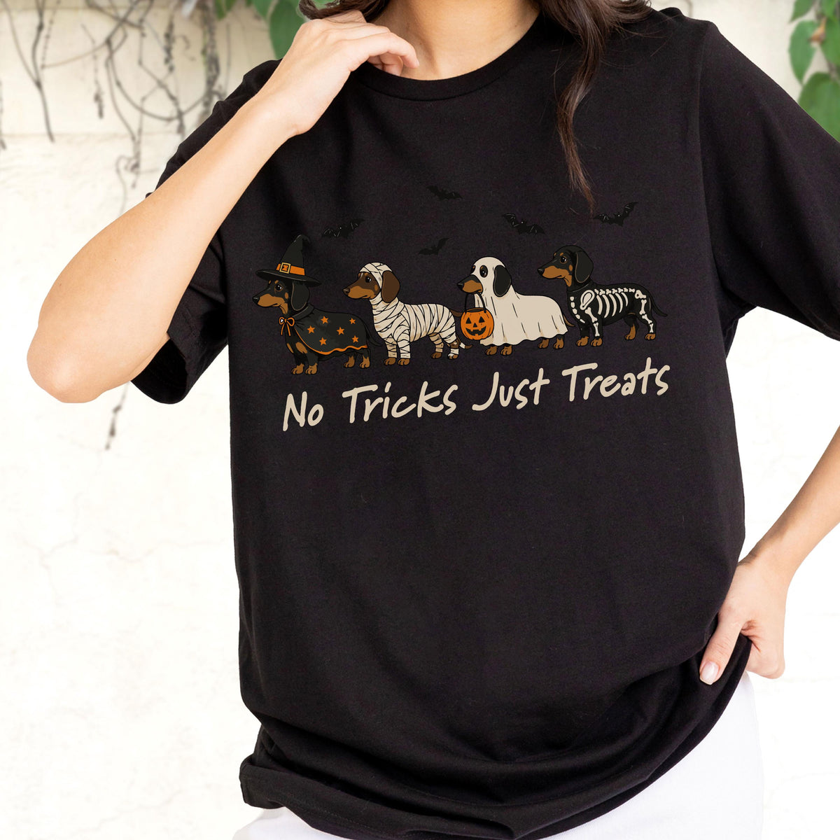 May include: A black t-shirt featuring a graphic of four dachshunds in Halloween costumes, including a witch, a mummy, a ghost holding a pumpkin bucket, and a skeleton. The text "No Tricks Just Treats" is printed in beige below the dogs.