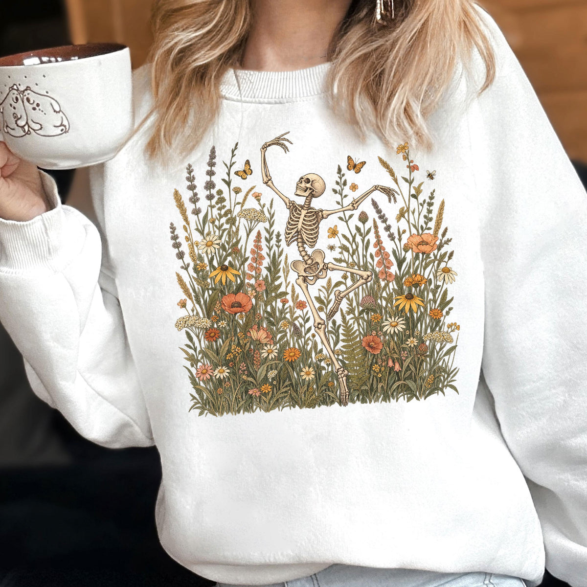 May include: A white crewneck sweatshirt featuring a graphic print of a skeleton dancing amidst a field of colorful wildflowers, tall grasses, and butterflies. The design includes various orange, yellow, and green floral elements. A white mug is also visible.