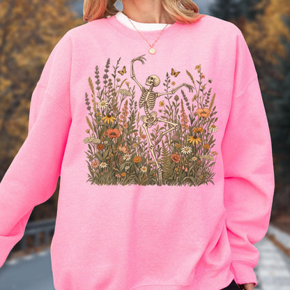 May include: A bright pink long-sleeve sweatshirt featuring a central graphic illustration of a skeleton dancing amidst a dense field of colorful wildflowers, tall grasses, and small butterflies in shades of orange, yellow, and muted green.