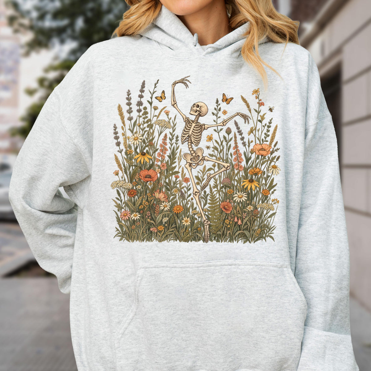 May include: A light gray heathered hooded sweatshirt featuring a central graphic print of a human skeleton dancing amidst a dense field of colorful wildflowers, tall grasses, and small butterflies in shades of orange, yellow, and muted green.