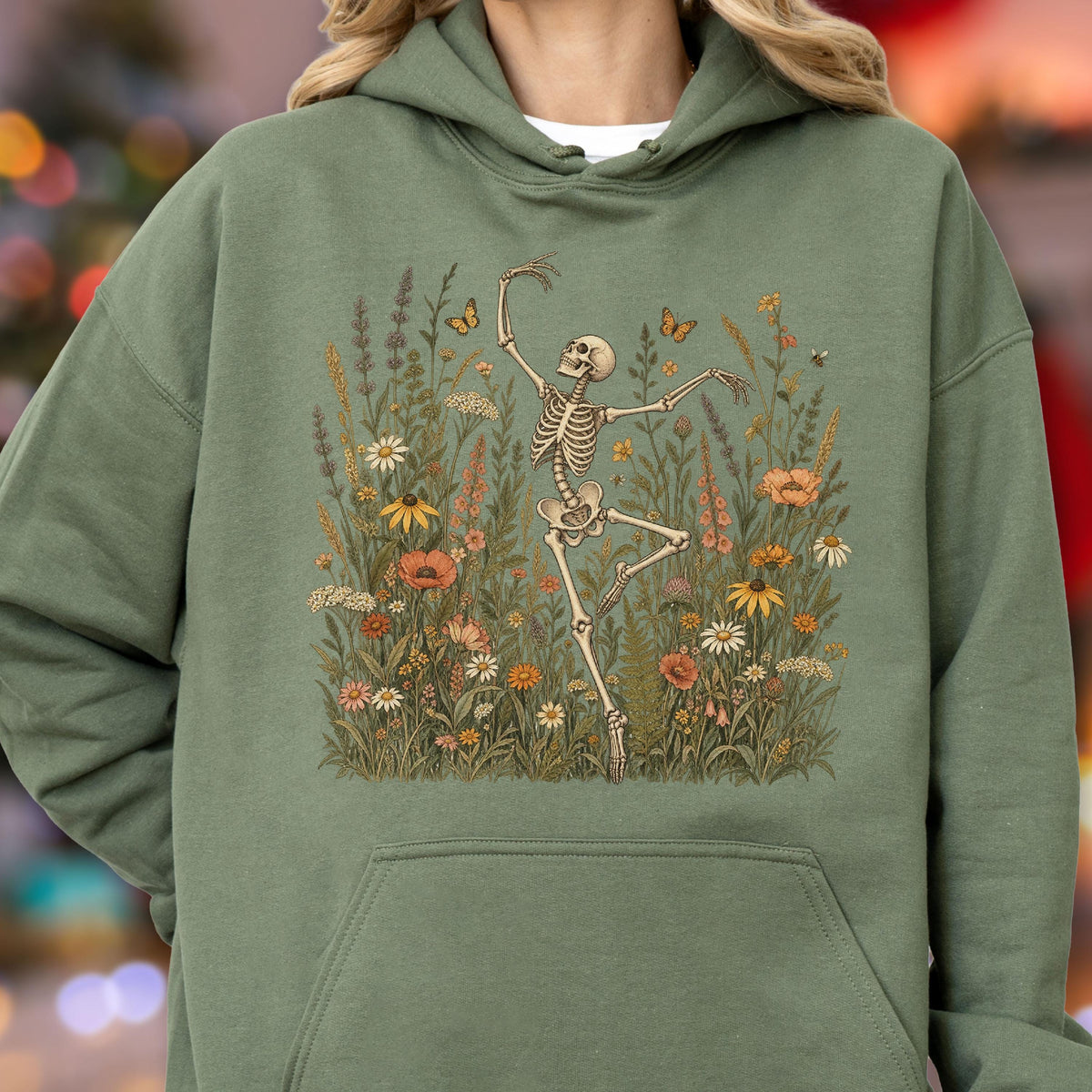 May include: An olive green hooded sweatshirt featuring a graphic print of a skeleton dancing amidst a field of colorful wildflowers and butterflies. The design includes various botanical elements in shades of orange, yellow, and white.
