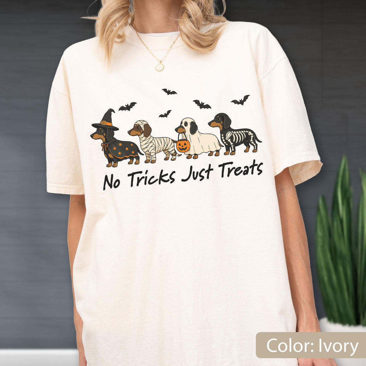 May include: An ivory t-shirt featuring a graphic of four dachshunds in Halloween costumes, including a witch, a mummy, a ghost holding a pumpkin bucket, and a skeleton. The text "No Tricks Just Treats" is printed below the dogs with small bats.