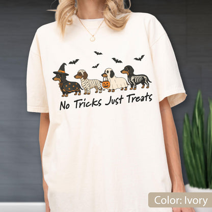 May include: An ivory t-shirt featuring a graphic of four dachshunds in Halloween costumes, including a witch, a mummy, a ghost holding a pumpkin bucket, and a skeleton. The text "No Tricks Just Treats" is printed below the dogs with small bats.