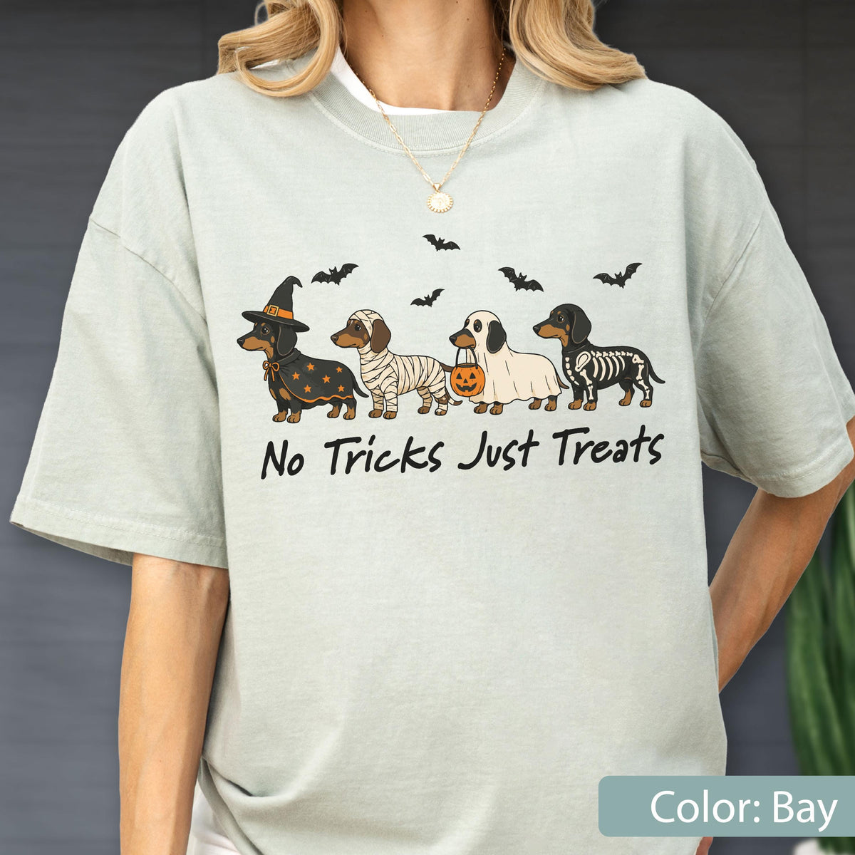 May include: A light sage green short-sleeve t-shirt featuring a graphic of four dachshunds in Halloween costumes, including a witch, a mummy, a ghost, and a skeleton, with bats and the text No Tricks Just Treats printed in black ink.