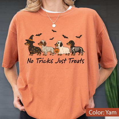 May include: A yam-colored t-shirt featuring a graphic of four dachshunds in Halloween costumes, including a witch, a mummy, a ghost, and a skeleton. Above the dogs are black bats, and below is the text No Tricks Just Treats in black.