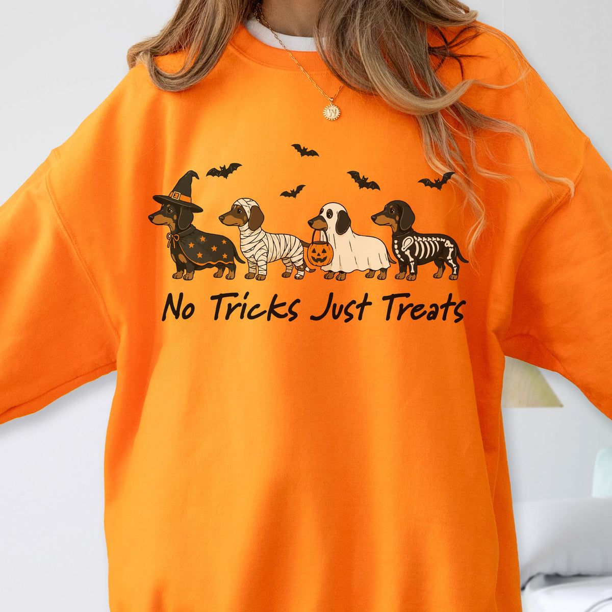 May include: An orange sweatshirt featuring a graphic of four dachshunds in Halloween costumes, including a witch, a mummy, a ghost holding a pumpkin bucket, and a skeleton. Above the dogs are black bats, and below is the text No Tricks Just Treats.