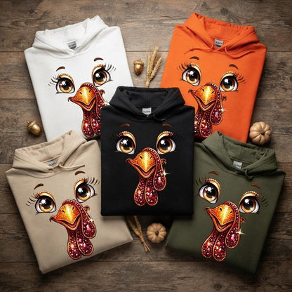 May include: Five hooded sweatshirts in white, orange, black, beige, and olive green. Each hoodie features a central graphic of a turkey face with large brown eyes, long eyelashes, and a textured, sparkling orange and red beak and wattle design.