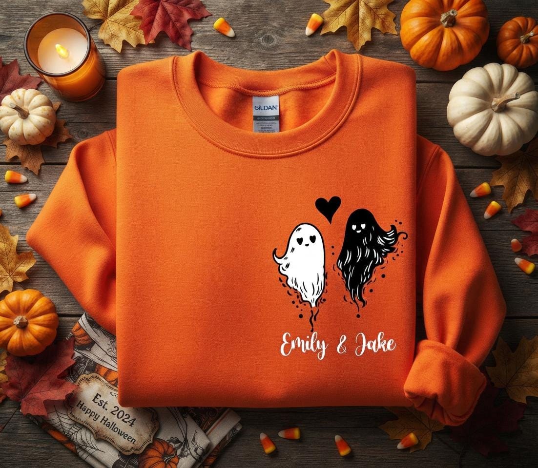 May include: An orange crewneck sweatshirt featuring a black graphic of two ghosts, one white and one black, with a small black heart between them. Below the ghosts, the text Emily & Jake is printed in a white script font on the front chest area.