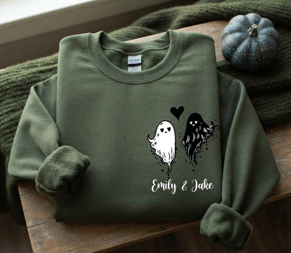 May include: An olive green crewneck sweatshirt featuring a graphic of two ghosts, one white and one black, with heart-shaped eyes. A small black heart is positioned between the ghosts, and the text "Emily & Jake" is printed below in white.