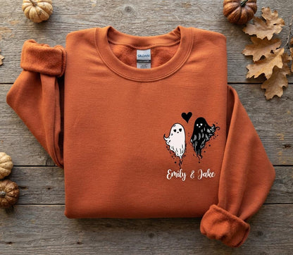 May include: A burnt orange crewneck sweatshirt featuring a graphic on the left chest. The design includes a white ghost and a black ghost with a small black heart between them. Below the ghosts, the text Emily & Jake is printed in white script.