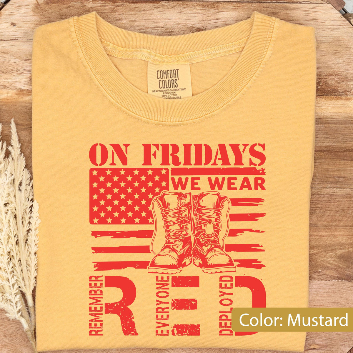 May include: A mustard yellow short-sleeve t-shirt featuring a red graphic print. The design includes a stylized American flag, a pair of military boots, and the text: ON FRIDAYS WE WEAR RED REMEMBER EVERYONE DEPLOYED. The shirt has a crew neckline.