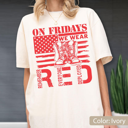 May include: An ivory t-shirt featuring a red graphic of the American flag, a pair of combat boots, and the text "ON FRIDAYS WE WEAR RED" along with "REMEMBER EVERYONE DEPLOYED" printed in a distressed, bold, block-style font across the chest.