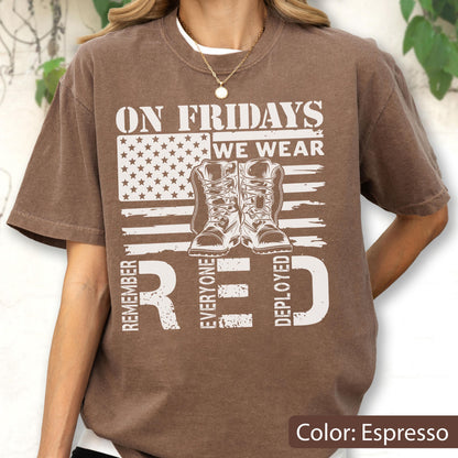May include: An espresso brown short-sleeve t-shirt featuring a white graphic print of an American flag, a pair of military boots, and the text ON FRIDAYS WE WEAR RED REMEMBER EVERYONE DEPLOYED. The shirt has a crew neckline and a relaxed fit.