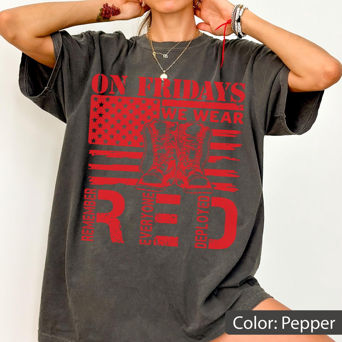 May include: A pepper-colored t-shirt featuring a red graphic of an American flag, a pair of combat boots, and the text: ON FRIDAYS WE WEAR RED, REMEMBER EVERYONE DEPLOYED. The design is centered on the chest of the short-sleeved, crew-neck garment.