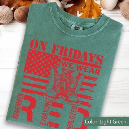 May include: A light green t-shirt featuring a red graphic print of an American flag, a pair of combat boots, and the text ON FRIDAYS WE WEAR REMEMBER EVERYONE DEPLOYED. The shirt has a crew neckline and a Comfort Colors brand label at the neck.