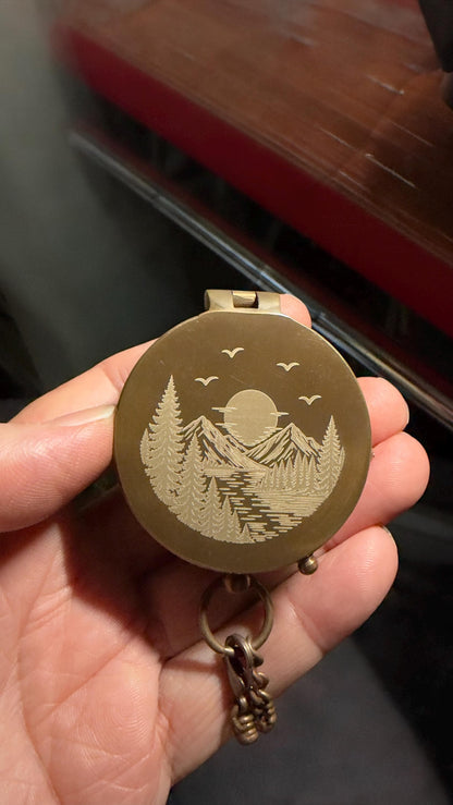 May include: A round, brass-colored metal pocket compass featuring a laser-engraved landscape design on its lid. The engraving depicts a sun rising over mountain peaks, a river flowing through a forest of pine trees, and three birds in the sky.