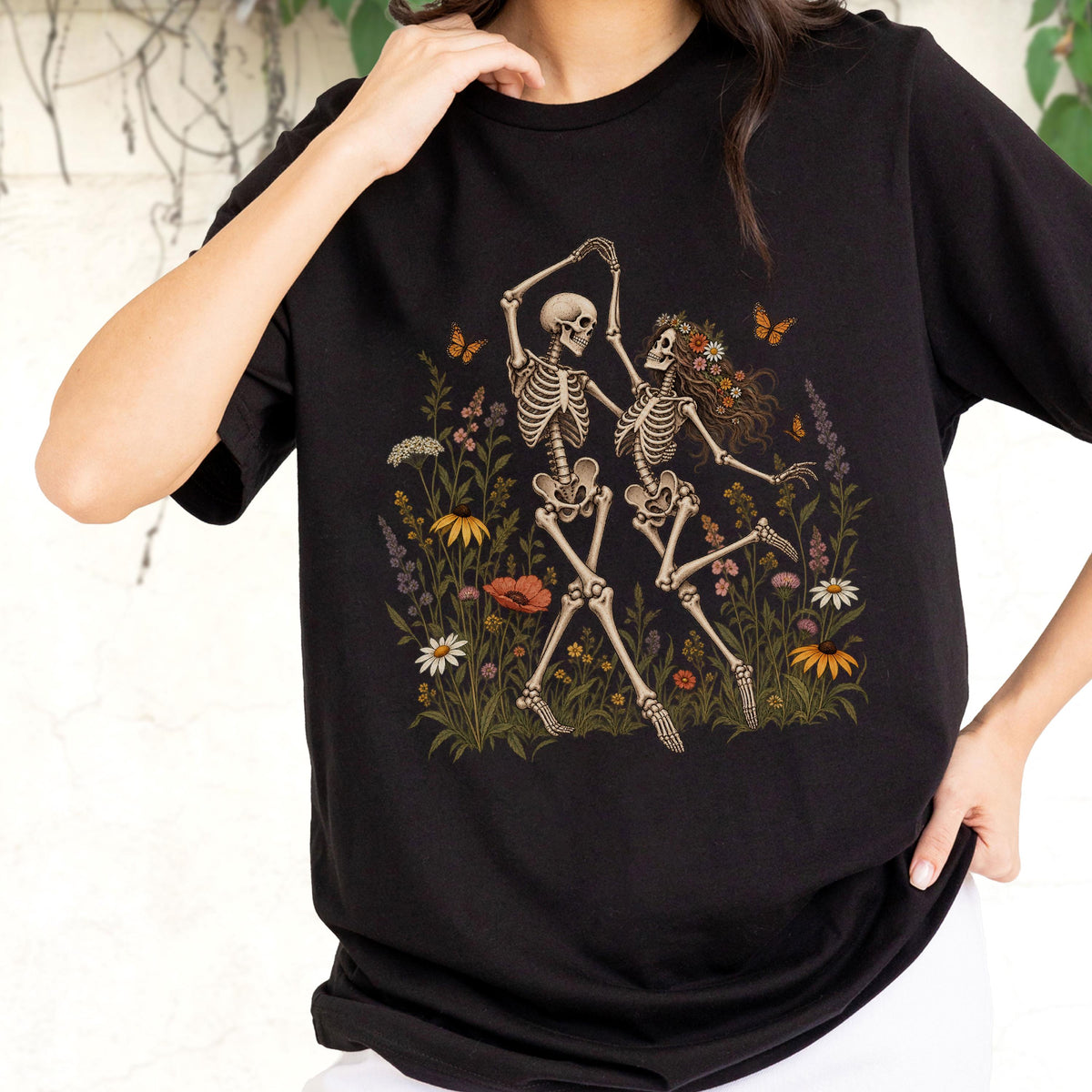 May include: A black t-shirt featuring a graphic print of two skeletons dancing in a field of colorful wildflowers. The skeletons are surrounded by various blossoms, green foliage, and small orange butterflies, creating a detailed scene across the chest.