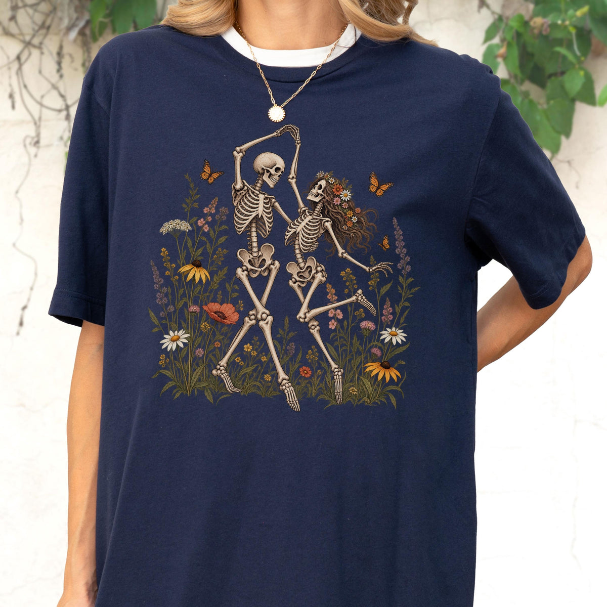 May include: A navy blue t-shirt featuring a graphic print of two skeletons dancing in a field of colorful wildflowers. The skeletons are surrounded by various blossoms, including daisies and poppies, with small orange butterflies fluttering nearby.