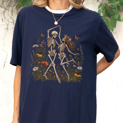 May include: A navy blue t-shirt featuring a graphic print of two skeletons dancing in a field of colorful wildflowers. The skeletons are surrounded by various blossoms, including daisies and poppies, with small orange butterflies fluttering nearby.