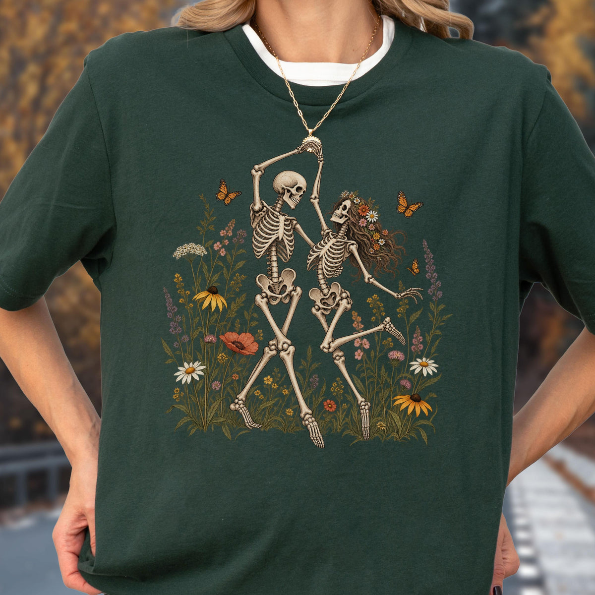 May include: A dark green short-sleeve t-shirt featuring a graphic print of two skeletons dancing in a field of colorful wildflowers and butterflies. The skeletons are depicted in a light beige tone, surrounded by various blossoms and orange butterflies.