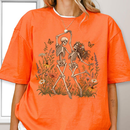 May include: A bright orange short-sleeve t-shirt featuring a graphic print of two skeletons dancing in a field of colorful wildflowers and butterflies. The skeletons are positioned in the center, surrounded by various floral elements and insects.