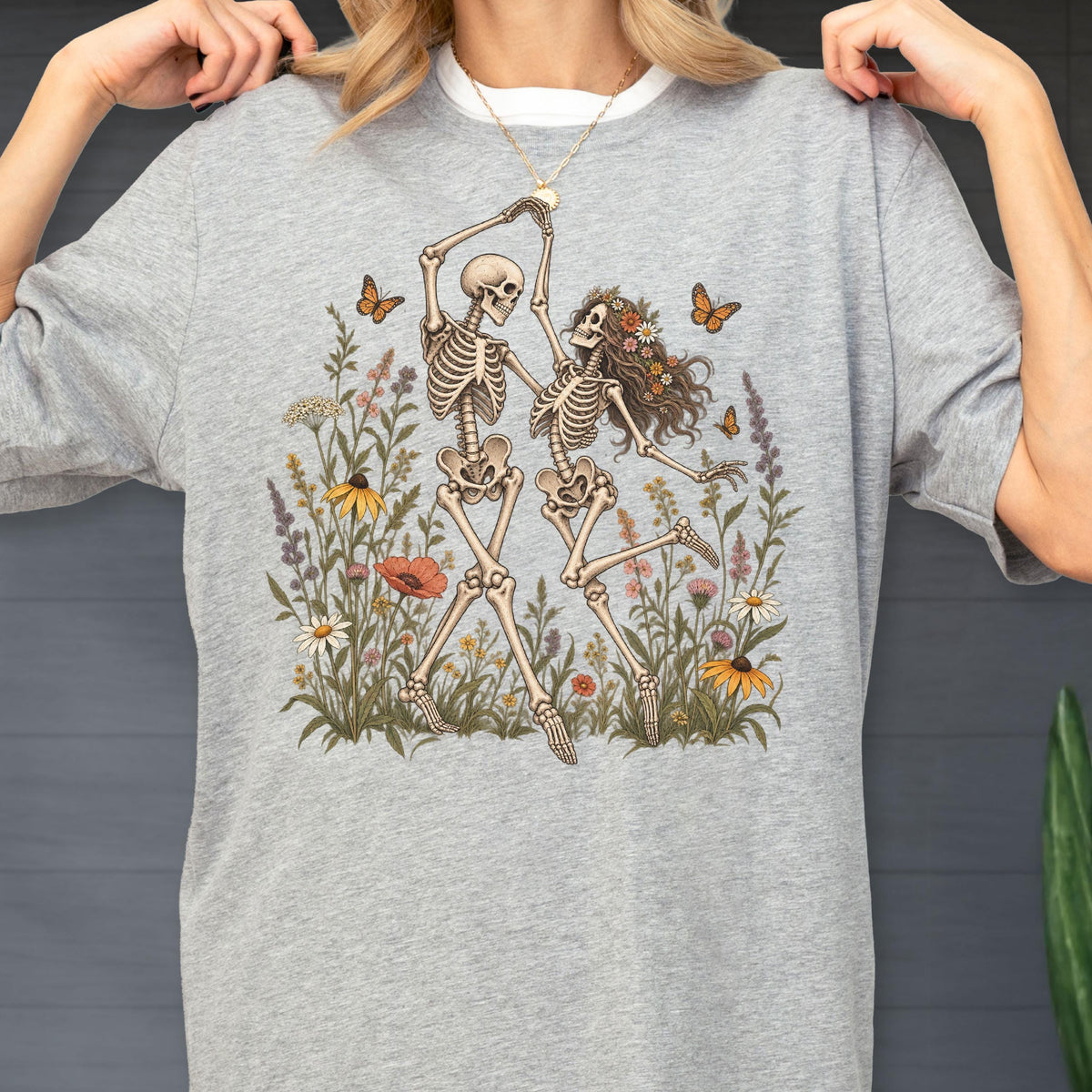 May include: A heather gray t-shirt featuring a graphic illustration of two skeletons dancing in a field of colorful wildflowers. The skeletons are surrounded by various blossoms and orange butterflies, with one skeleton wearing a crown of flowers.