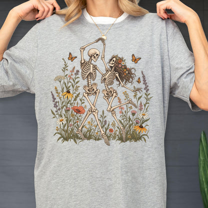 May include: A heather gray t-shirt featuring a graphic illustration of two skeletons dancing in a field of colorful wildflowers. The skeletons are surrounded by various blossoms and orange butterflies, with one skeleton wearing a crown of flowers.