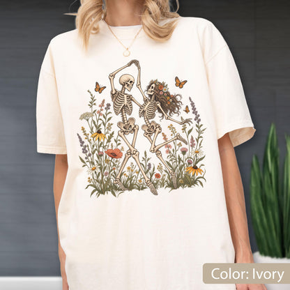 May include: An ivory t-shirt featuring a graphic print of two skeletons dancing in a field of colorful wildflowers. The skeletons are surrounded by various blossoms and small orange butterflies. The design is centered on the front of the shirt.