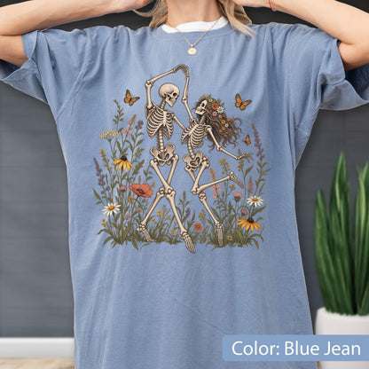 May include: A blue jean colored t-shirt featuring a graphic print of two skeletons dancing in a field of colorful wildflowers. The design includes orange butterflies, various green plants, and flowers in shades of yellow, white, and red, centered on the chest.