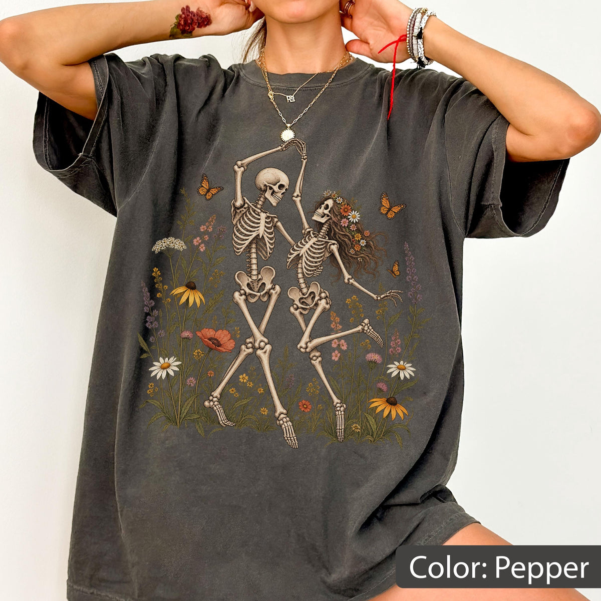 May include: A charcoal gray t-shirt featuring a graphic print of two skeletons dancing amidst a field of colorful wildflowers and butterflies. The shirt has a crew neckline, short sleeves, and a relaxed fit, showcasing a detailed illustration.