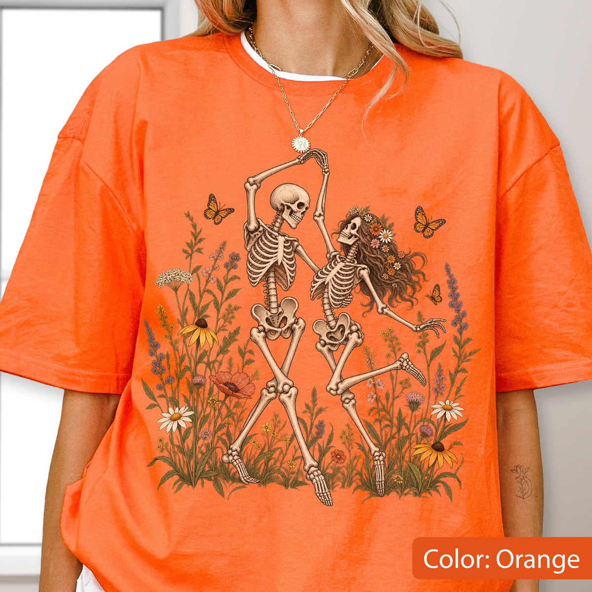 May include: An orange t-shirt featuring a graphic print of two skeletons dancing in a field of colorful wildflowers and butterflies. The skeletons are positioned in the center of the shirt, surrounded by various botanical elements and insects.