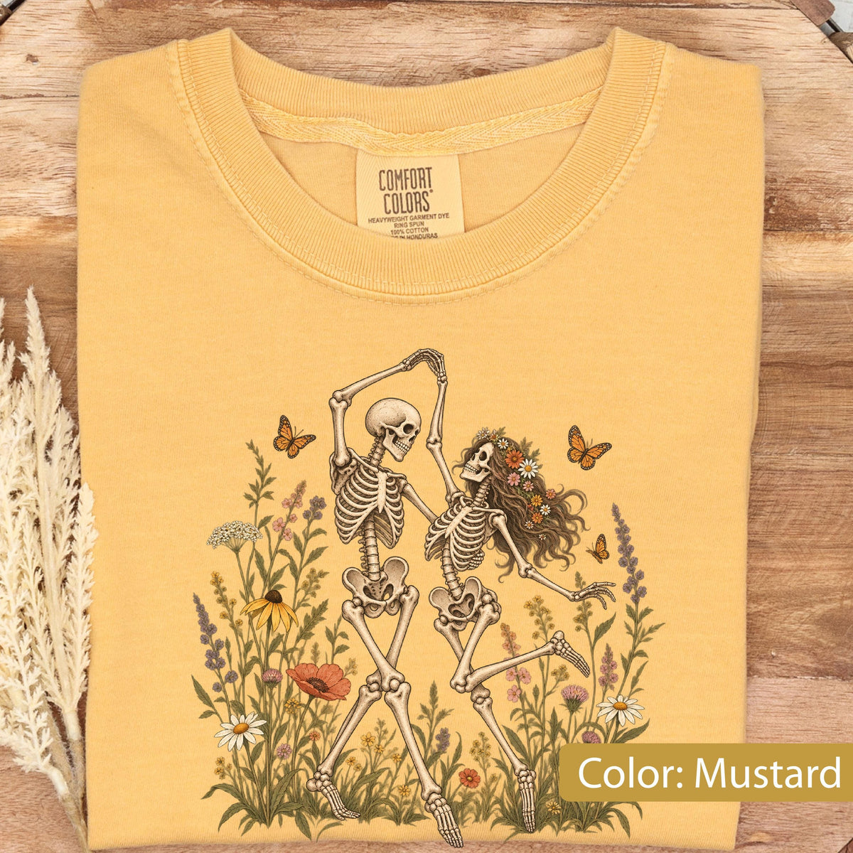 May include: A mustard yellow t-shirt featuring a graphic print of two skeletons dancing amidst a field of colorful wildflowers and butterflies. The inner neck label displays the text Comfort Colors, Heavyweight Garment Dye, Ring Spun, 100% Cotton.