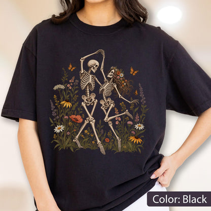 May include: A black t-shirt featuring a graphic print of two skeletons dancing in a field of colorful wildflowers. The skeletons are surrounded by various blossoms, green foliage, and small orange butterflies, creating a detailed botanical scene.