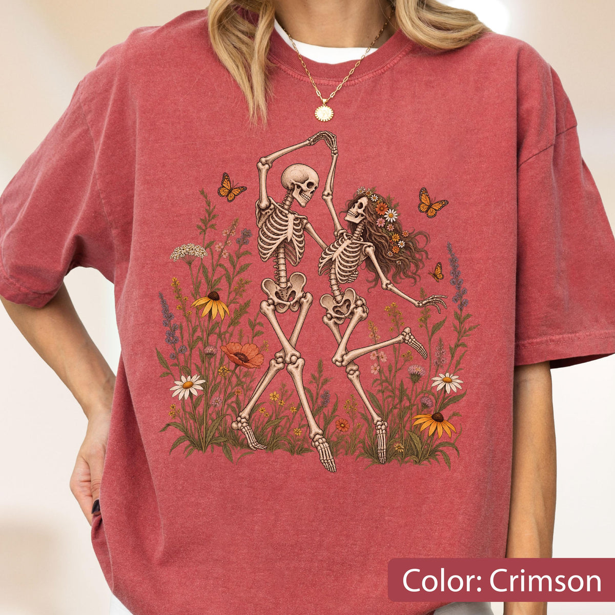 May include: A crimson short-sleeve t-shirt featuring a graphic print of two skeletons dancing in a field of colorful wildflowers and butterflies. The design includes various floral elements in shades of yellow, orange, and white, with orange butterflies.