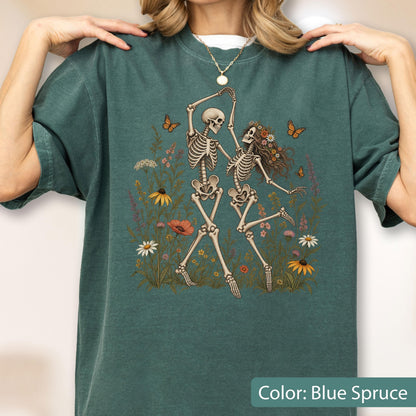May include: A blue spruce t-shirt featuring a graphic print of two skeletons dancing in a field of colorful wildflowers and butterflies. The design includes various floral elements in shades of orange, yellow, and white, set against the dark fabric.