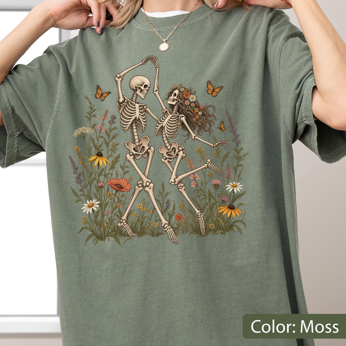 May include: A moss green t-shirt featuring a graphic print of two skeletons dancing in a field of wildflowers. One skeleton has long hair adorned with flowers. Several orange butterflies are depicted flying around the skeletons and flowers.
