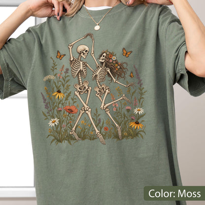 May include: A moss green t-shirt featuring a graphic print of two skeletons dancing in a field of wildflowers. One skeleton has long hair adorned with flowers. Several orange butterflies are depicted flying around the skeletons and flowers.